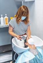 Hairdresser with face mask washing hair in the hot sink to the client. Safety measures for hairdressers in the Covid-19 pandemic. New normal