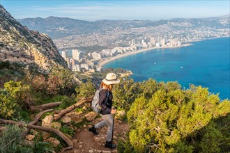 A young hiker wearing a hat on the descent path of the Penon de Ifach Natural Park with the city of Calpe in the background