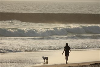 A woman walking with her little dog on the beach at sunset. Mid shot