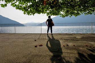 Woman with Shadow Standing Below a Beautiful Tree with Branch on the Waterfront with Railing to Lake Lugano with Mountain in a Sunny Summer Day in Bissone