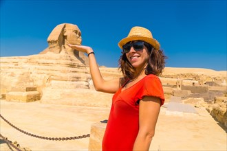 A young tourist in a red dress joking at the Great Sphinx of Giza and in the background the Pyramids of Giza