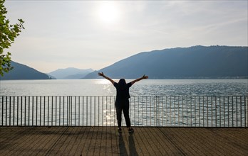 Woman with Shadow Standing with Arms Outstretched on the Waterfront with Railing to Lake Lugano with Mountain in a Sunny Summer Day in Bissone