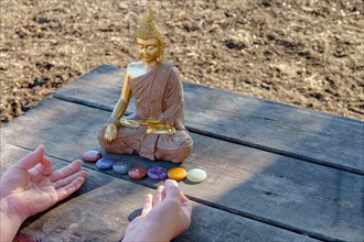 Woman's hands meditating in front of a Buddha statue with colorful chakra stones on a wooden table in a park in the sunshine