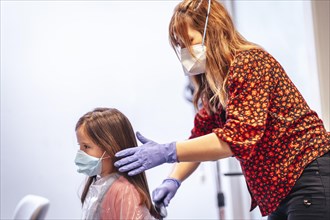 Hairdresser with mask and gloves combing the straight hair of a blonde girl