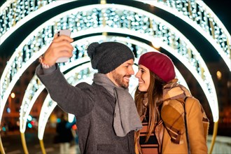 Winter portrait of a Caucasian couple enjoying the Christmas lights of the city taking a selfie with the mobile for social networks