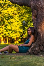 A young pretty Latina brunette with long straight hair leaning against a tree in a green dress. Sitting by a tree in the park