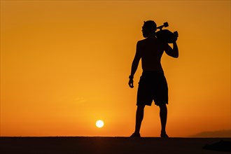 Silhouette of a free man holding a skateboard on his shoulder at bright sunset. Mid shot