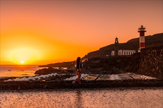 A young woman walking in the orange sunset in the salina and in the background the Fuencaliente Lighthouse on the route of the volcanoes south of the island of La Palma