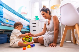 Young Caucasian mother playing with her in the room with toys. Baby less than a year learning the first lessons of her mother. Mother playing with her son playing sitting on the floor