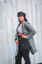 A young brunette Latina with a leather cap and a gray blazer in a portrait with a gray city background