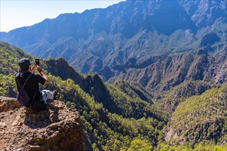 A young man resting after trekking at the top of La Cumbrecita sitting in the natural viewpoint and taking a photo with the mobile