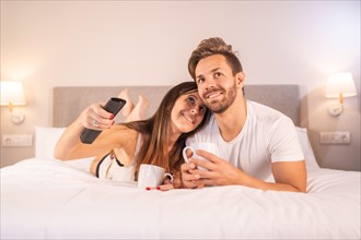 A couple in love watching TV on top of the hotel bed in pajamas