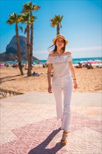 A Caucasian redhead dressed in white and with a straw hat walking along the beach of Calpe in summer