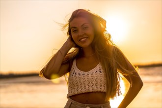 Summer lifestyle. A young blonde Caucasian woman in a white short wool sweater on a beach sunset. Smiling and looking at camera