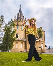 Blonde woman in a yellow vintage outfit with a hat walking next to a castle