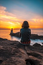 A young woman sitting on some rocks with a leather jacket in the orange sunset of the Pasajes San Juan lighthouse. Gipuzkoa