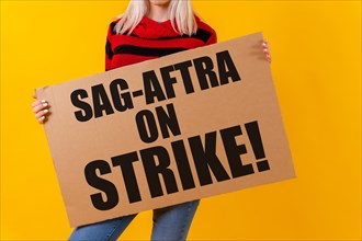A female actress holding a sign with the hollywood actors and writers strike slogan on a yellow background