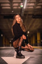 Caucasian brunette model with black jacket posing on the white arrow of the ground in an empty underground car park. Night urban session in the city