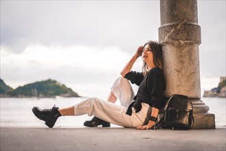 Lifestyle in the city with a blonde girl in white pants and black wool sweater near the beach. Sitting on the shallows of a beach at sunset