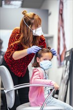 Hairdresser with mask and gloves finishing the pigtails to a girl. Reopening with security measures for hairdressers in the Covid-19 pandemic