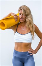 Fitness and yoga session with a young blonde Caucasian woman dressed in a casual outfit. Smiling on a white background with a mat on his shoulder