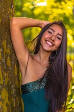 A young brunette Latina with long straight hair leaning against a tree in a green dress. Portrait of pensive and sensual young woman