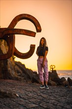 A young and pretty brunette Latina with long straight hair in a short black T-shirt and pink pants. Next to a sculpture of San Sebatian called Peine del Viento at sunset