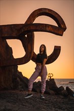 A young pretty brunette Latina with long straight hair in a short black T-shirt and pink pants. Perched next to a famous sculpture of Saint Sebatian called Peine del Viento