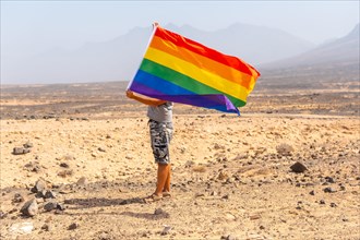 An unrecognizable gay person in a gray t-shirt and black cap with the LGBT flag waving in a desert with the wind