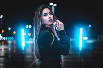 Caucasian brunette in pink wool jacket in an empty car park illuminated with blue light. Night urban session in the city