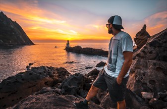 A relaxed young man in a cap looking at the orange sunset of the Pasajes San Juan lighthouse