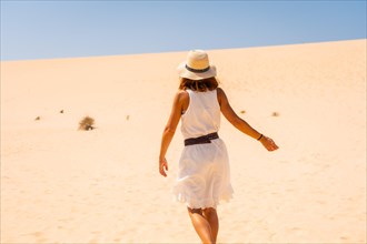 A young tourist in a white dress and a hat walking through the dunes of the Corralejo Natural Park