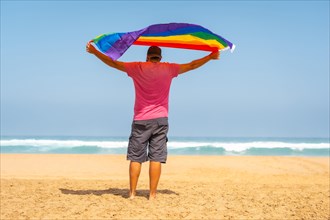 A gay person with a pink t-shirt and black cap with the LGBT flag on a beach