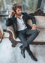 Street style with a young brunette caucasian man in a black suit and a white shirt in a luxurious hotel. Sitting on a traditional sofa looking at camera