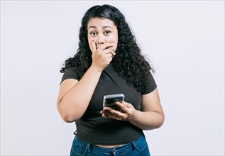 Surprised young woman covering mouth holding phone isolated. Astonished young girl holding phone and covering mouth isolated