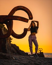 A young pretty brunette Latina with long straight hair in a short black T-shirt and pink pants. Orange sunset next to a famous sculpture of San Sebatian called Peine del Viento