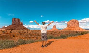Man in white t-shirt and green hat with arms raised in Monument Valley National Park at The Mittens and Merrick Butte point. Utah