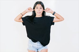 Displeased teen girl covering ears and sticking out tongue isolated. Latin girl covering her ears and making faces isolated