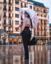 Posing of a young brunette Latina with a leather hat and black pants in the autumn rain with a transparent umbrella in the city
