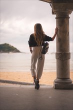 Lifestyle in the city with a blonde girl in white pants and a leather jacket near the beach. Photos next to a column on the beach