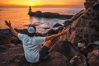 A young man sitting on some rocks watching the orange sunset of the Pasajes San Juan lighthouse