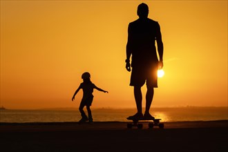 Silhouette of a man and a child skating on skateboard and roller skates at bright sunset. Mid shot