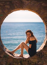 A young pretty red-haired Caucasian girl smiling sitting in a blue dress by the sea