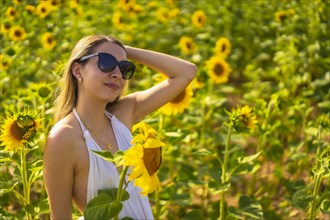 A blonde Caucasian woman with sunglasses and a white dress in a beautiful field of sunflowers on a summer afternoon