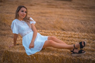 A young blonde Caucasian woman in a white dress in a field of dry straw atop a haystack. In a dry cultivated field in Navarra