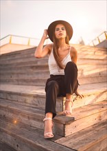 Young brunette in a black hat and white t-shirt enjoying the summer sitting on some wooden stairs by the sea