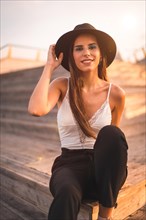 Portrait of a young brunette in a black hat and white t-shirt enjoying the summer sitting on some wooden stairs by the sea