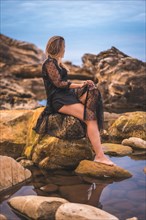 Summer lifestyle with a young brunette Caucasian woman in a long black transparent dress on some rocks near the sea on a summer afternoon. Sitting on top of a rock in a natural pool