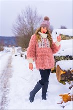 Posing blonde model with pink fur jacket and purple hat in the snow. Next to some ice-cut trees on a footpath