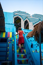 A young woman in a red dress climbing some colorful stairs that go up to a beautiful terrace of a traditional blue house in a Nubian village next to the Nile river and near the city Aswan. Egypt